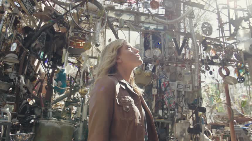 A blonde woman wearing a brown leather jacket enters a structure with cluttered metallic objects lining the wall