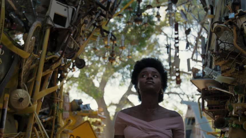 A young woman walks through a narrow alley filled with cluttered objects in a natural setting