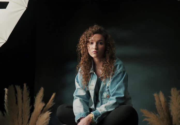 A young woman with curly hair sits cross legged on a wooden box wearing a denim jacket and looking forward