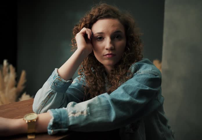 A young woman sits in a room and drops her hand down from her face to her other wrist