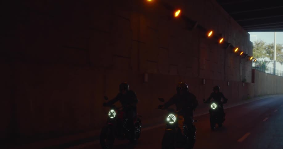 Three bikers with their motorcycle headlights on are riding on an asphalt street passing under a tunnel