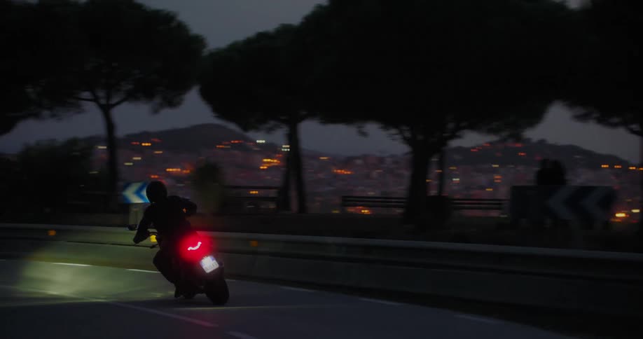A motorcyclist riding on a road at dusk with city lights in the background and trees lining the road
