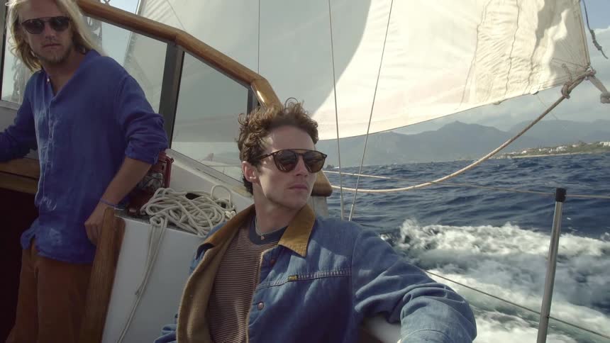 Two men are in a sailboat that is sailing on a sea and the wind is blowing through their hair