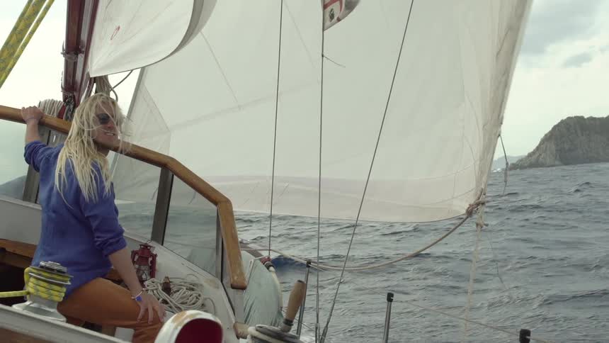 Back view of a woman on a sailboat with a large white sail unfurled navigating through open water with rocky coastlines in the distance
