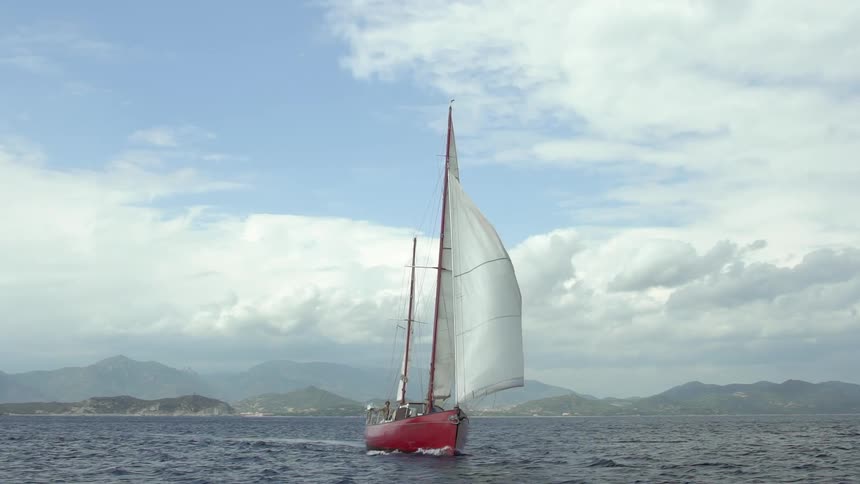A red sailboat sailing across choppy water in a wide open area with mountains in the background