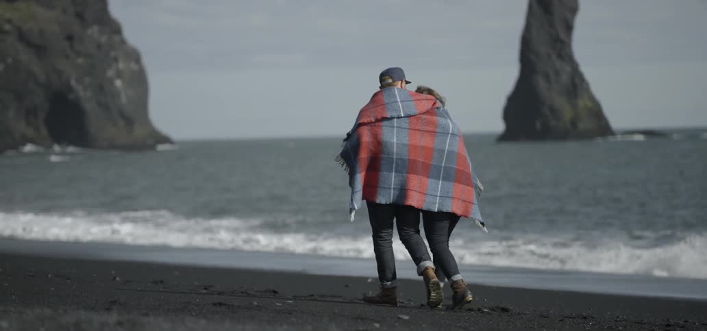 A man and woman wrapped in a blanket stroll on a beach during the day