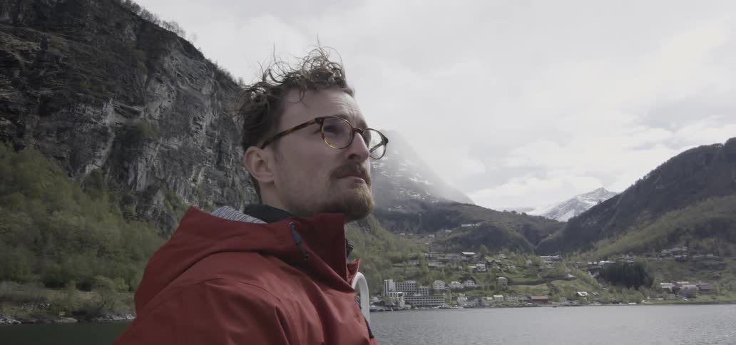 A man in glasses looks around near a body of water