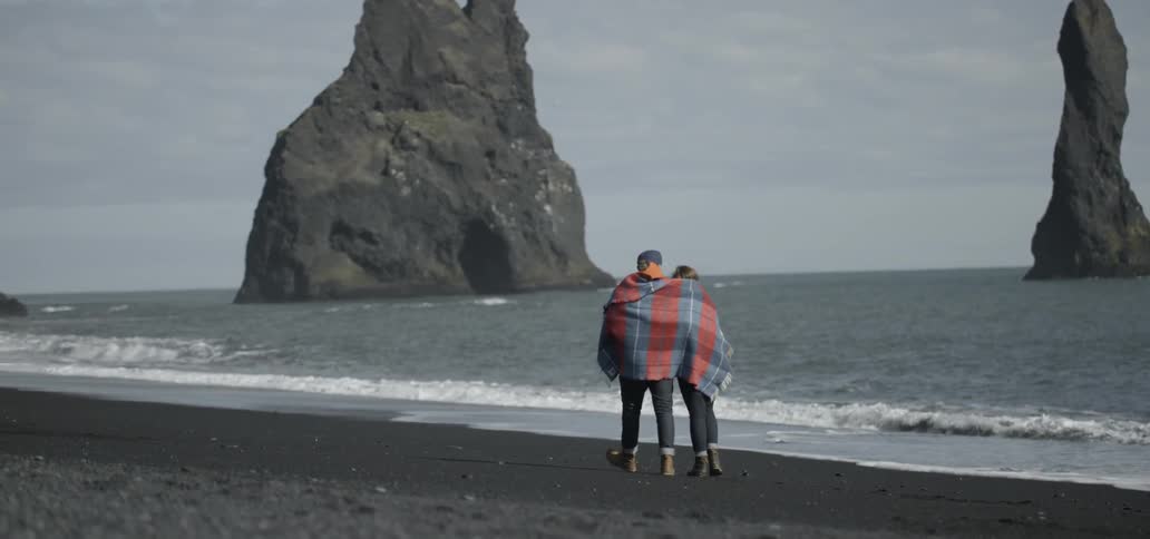 Couple walking along the shore while sharing a blanket around their torsos on a cloudy day