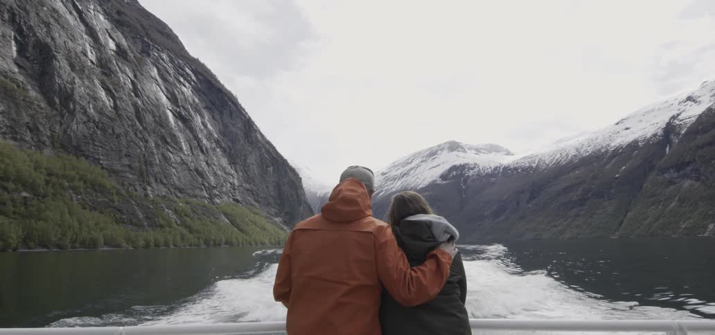 A couple on a boat deck looks out at the water and mountains
