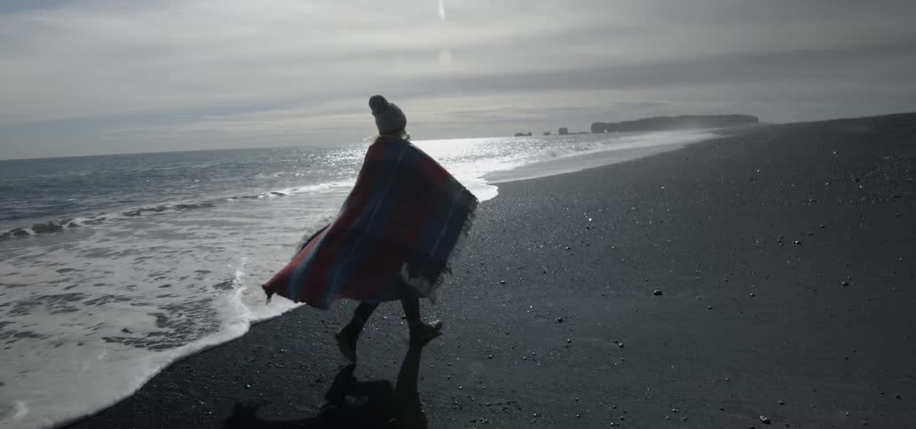 A woman wearing a blanket walks down the beach