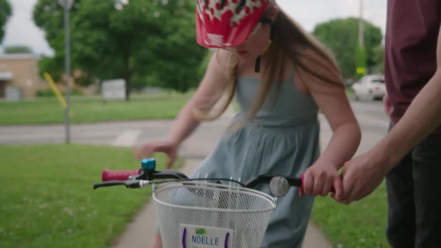 A young girl wearing a polka dot helmet getting onto a bicycle with the help of her dad on a sidewalk