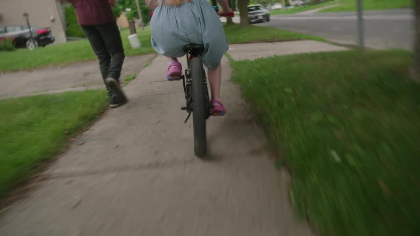 A young girl riding a bike on a sidewalk as her father runs next to her