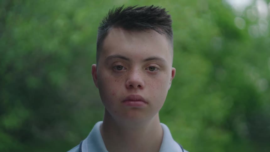 The face of a young man staring straight ahead and behind him are some blurred green leaves
