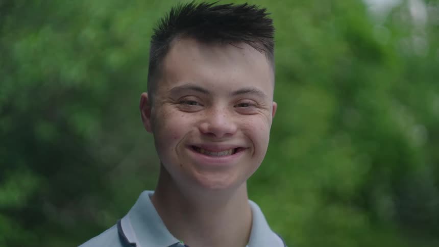 A young man looking forward is smiling and behind him there are some blurred trees