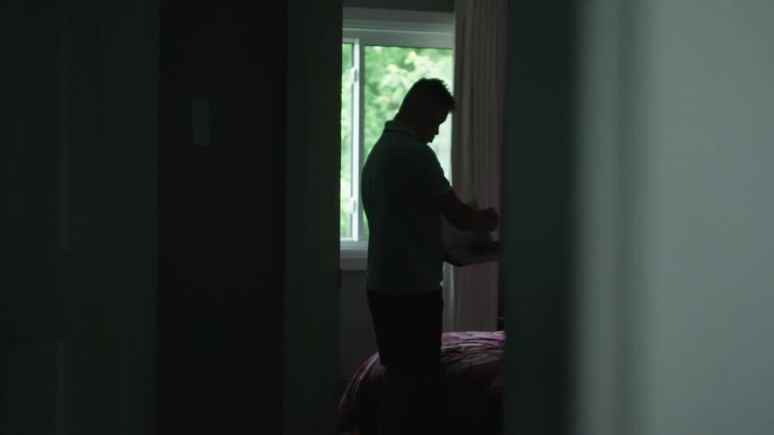 The silhouette of a man standing in his bedroom at the foot of his bed looking contemplatively
