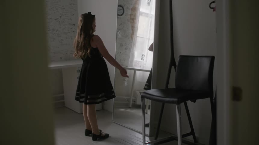 A young girl with Down syndrome in a dark dress stands in front of a mirror in a dimly lit room