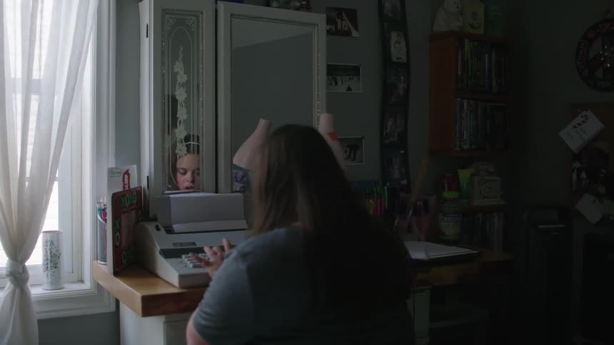 The backside of a young girl typing on a typewriter on a desk in her bedroom near a window