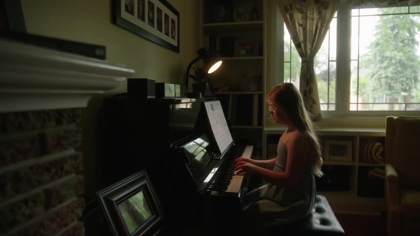 A young girl playing piano lit up by a small lamp in a living room on a bright sunny day