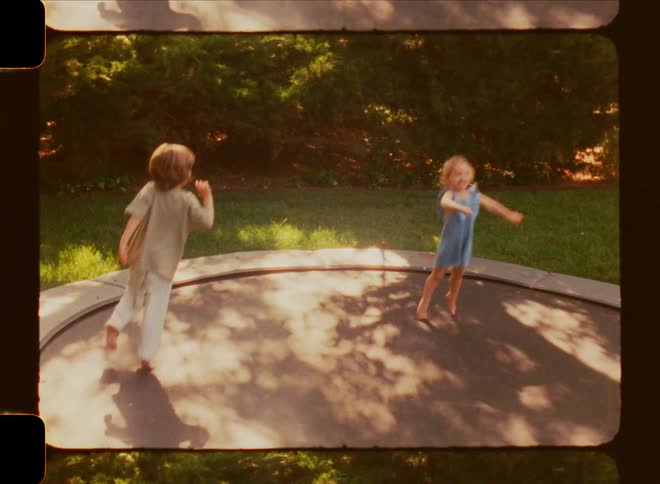 Two young children jumping up and down and running around on a trampoline in the middle of a summer day