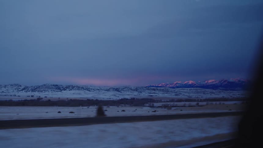Snow covered mountains in the distance past a highway with cars and trucks traveling on it at dusk