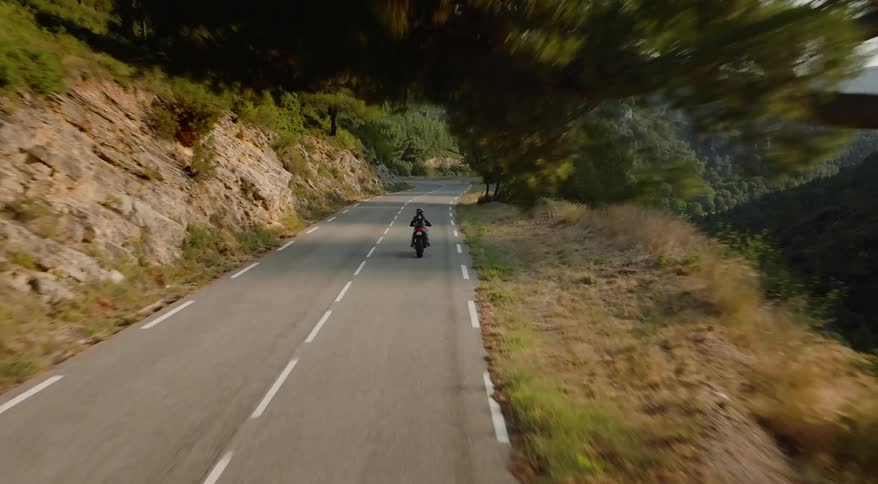 A person riding a motorcycle on an asphalt street with white stripes and in the middle of a region with mountains and trees