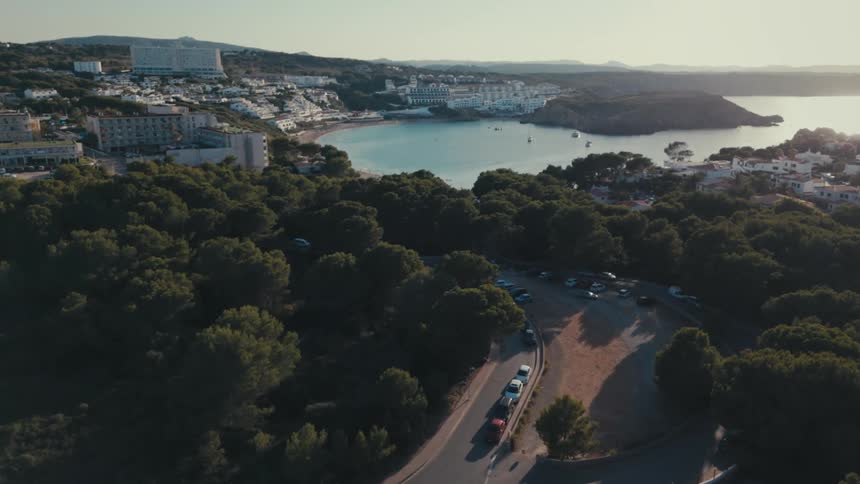 An aerial view of an asphalt street with several trees around it and further ahead a city facing the sea