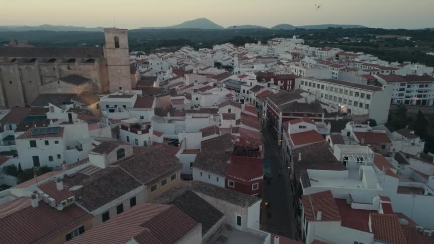 An aerial view of a city with white houses with orange roofs and some birds flying over the place