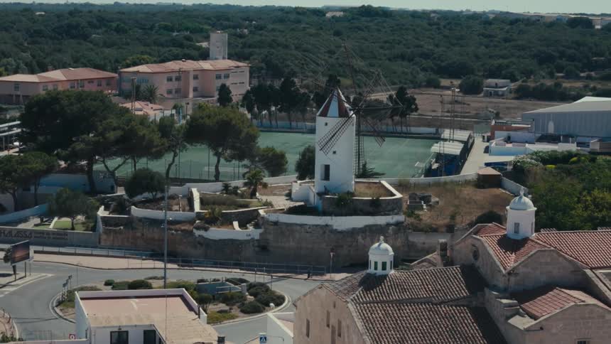 An aerial of a windmill in a small town with greek vibes surrounded by dense forests