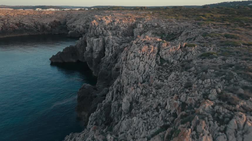 Waves flow against the base of a craggy cliff topped by the uneven terrain bordering a forest