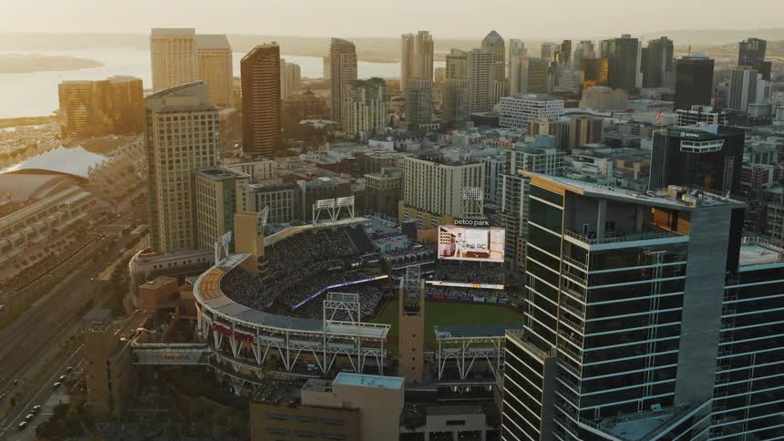An aerial of a crowded stadium in the middle of a city with buildings all around it and the sun setting on the horizon