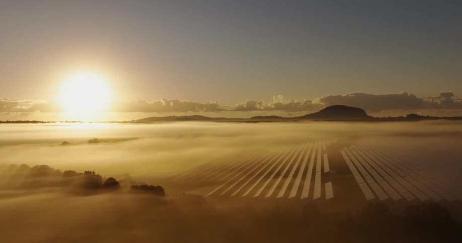 Bright sun shines from the horizon onto a solar farm located in a fog shrouded landscape