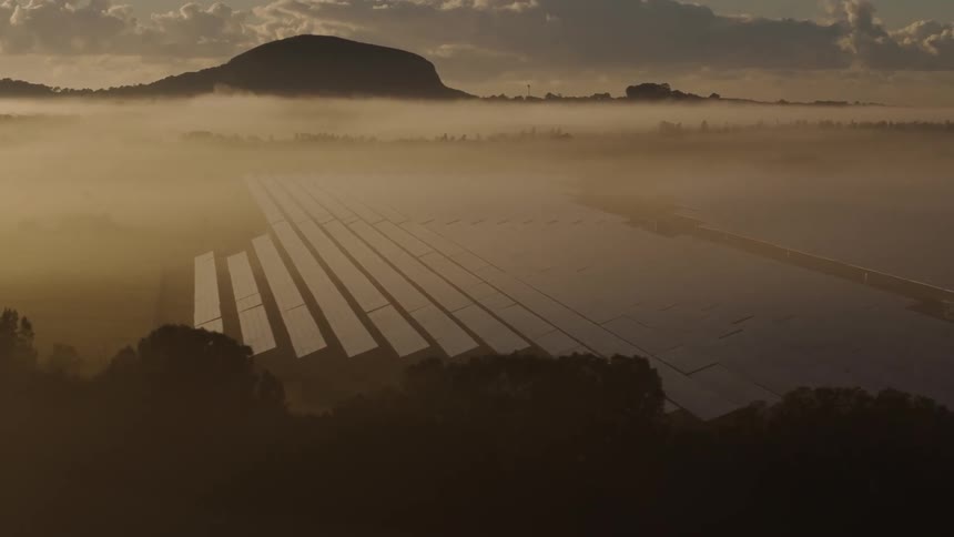 Dense fog covers solar panels in a field bracketed by trees and a silhouetted hill on a cloudy day