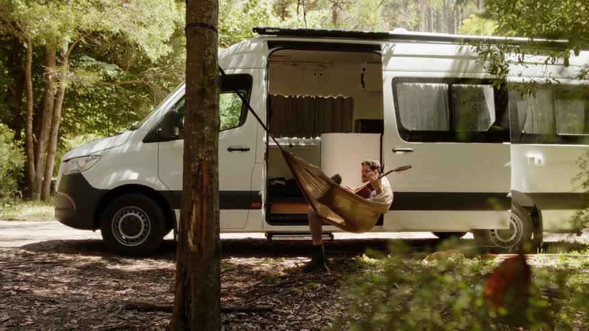 A man lying in a hammock playing the guitar and there is a trailer behind him