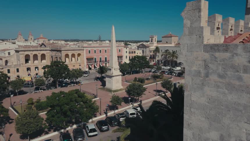 A city square with an obelisk in the center and several trees and cars parked around it