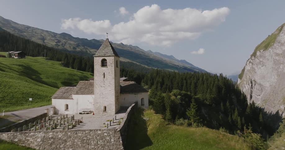 An aerial view of a small church situated on a grassy hill surrounded by mountains
