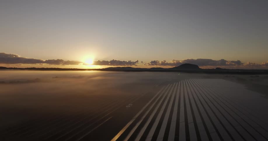 Setting sun shines onto rows of solar panels partially covered by fog
