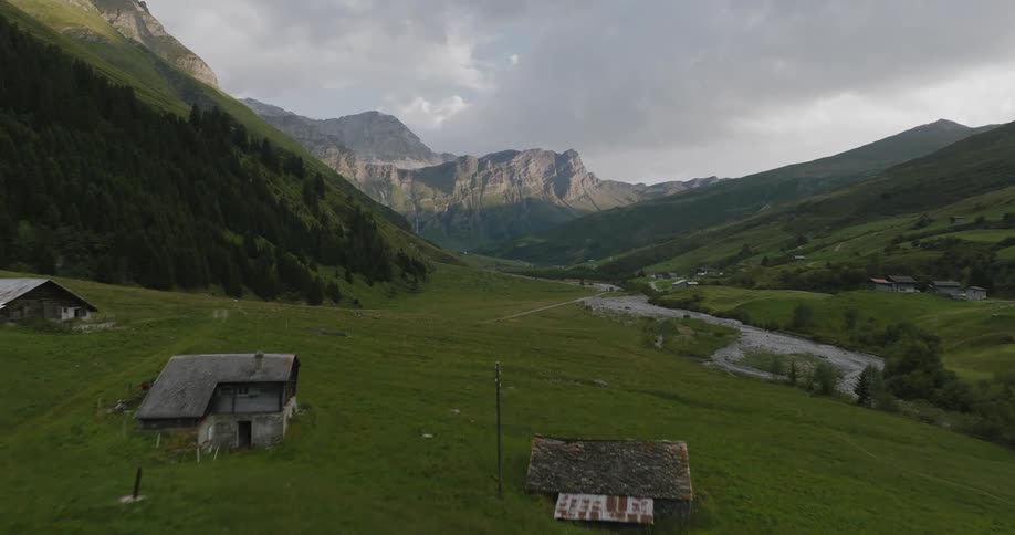 A landscape of a large grass field with some trees and mountains in the background and there are some wooden huts on the grass