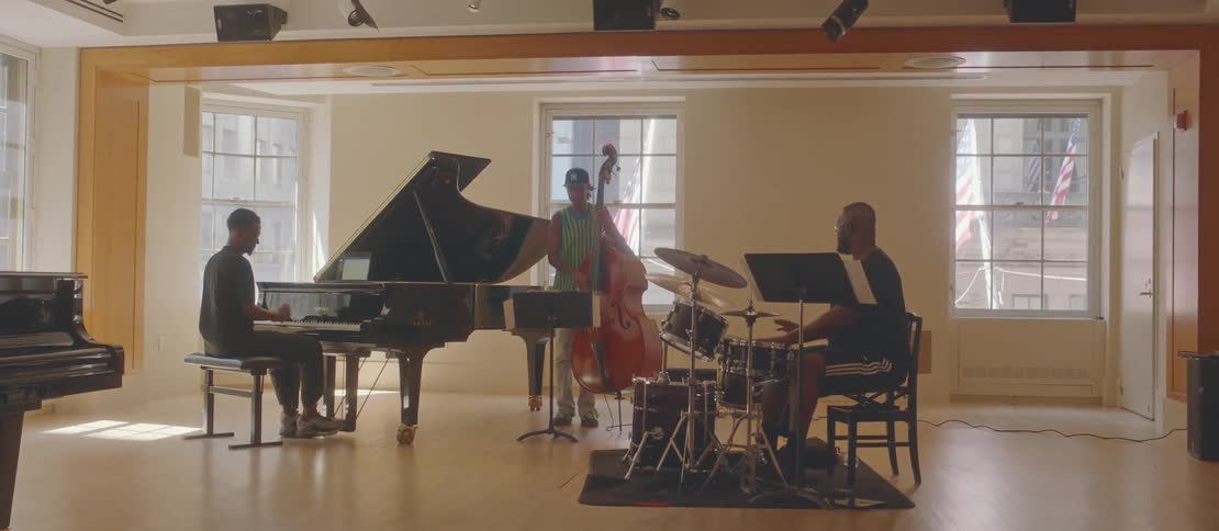 Three musicians in a welllit room with large windows playing a grand piano drums and upright bass