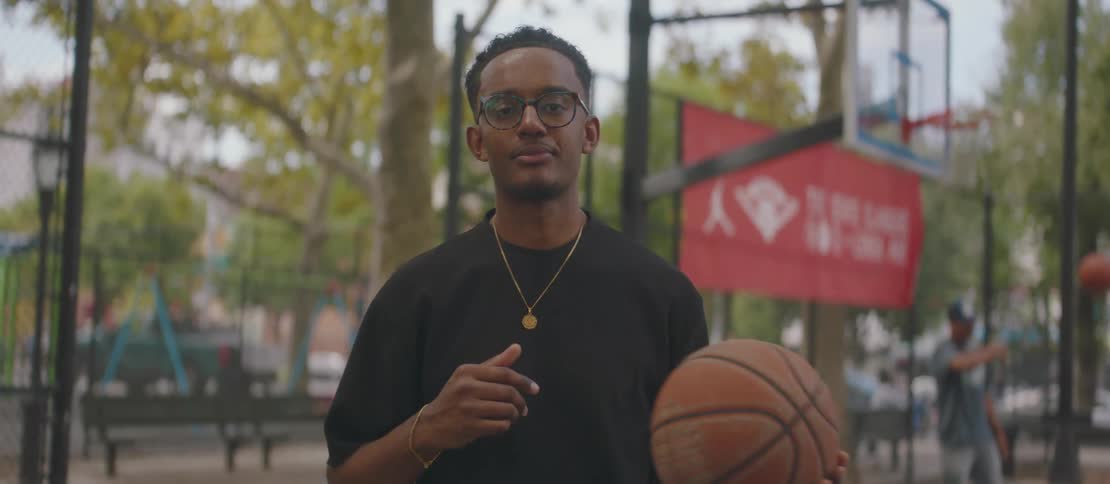 A black teenager stands on a basketball court smiling as he tosses a basketball from one hand to another