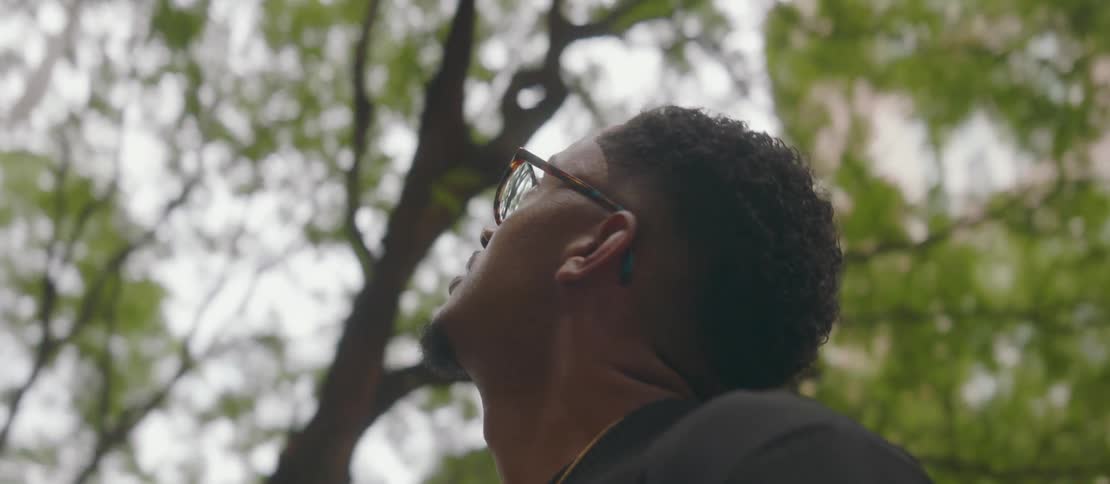 A young man wearing glasses is standing looking up in front of him at an apartment building with external stairs