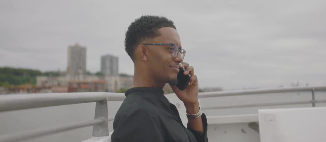 Man sitting near the rail on a ship cruising off the shore near a city answers the phone