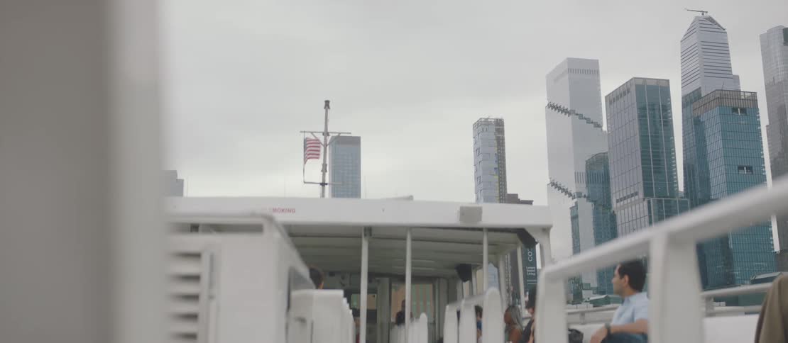 An american flag blows in the wind on top of a boat moving towards a downtown city area
