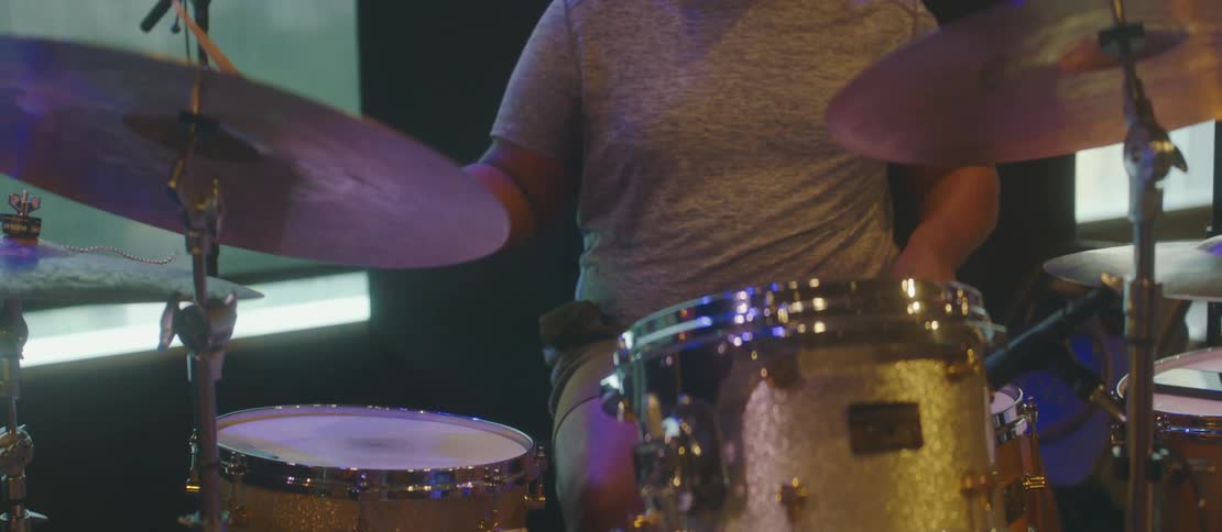 A male drummer is sitting while using his drumsticks to hit the drums