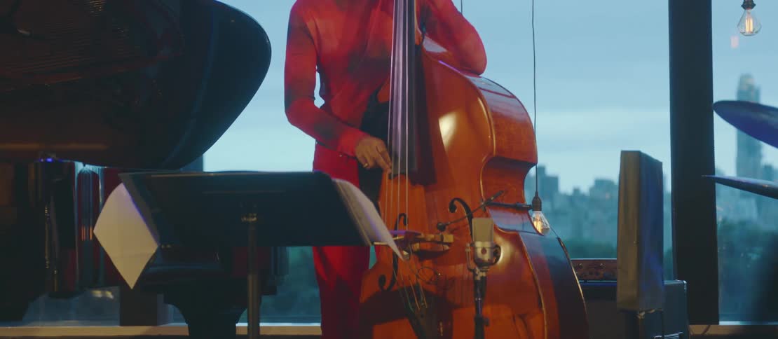 A man in red clothes is standing playing a big cielo violin and he is looking down