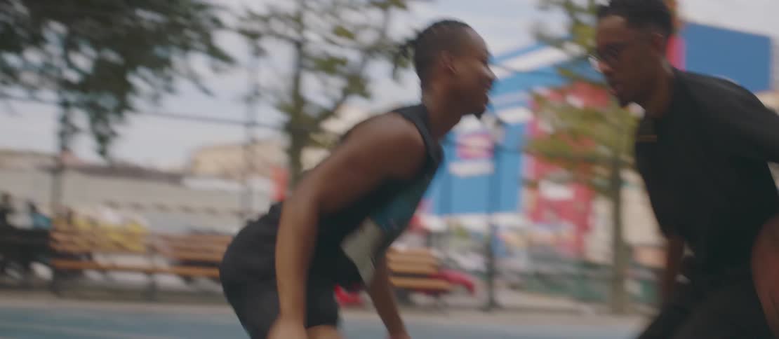 Two men playing basketball on a concrete court and one of them is going towards the other with the ball while bouncing it on the ground