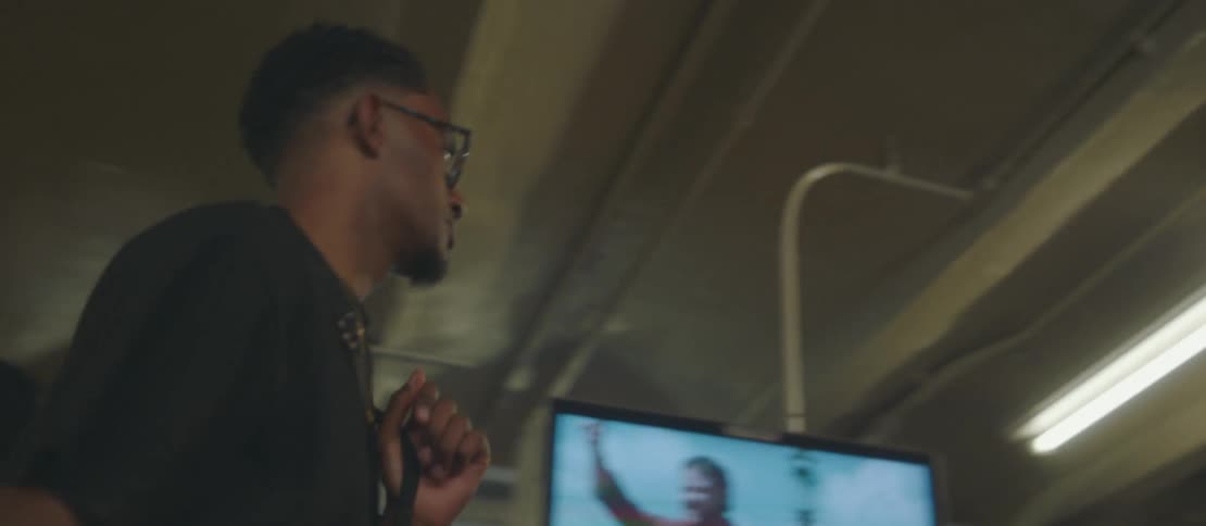 A young man wearing reading glasses is walking in a terminal with some people and some screens