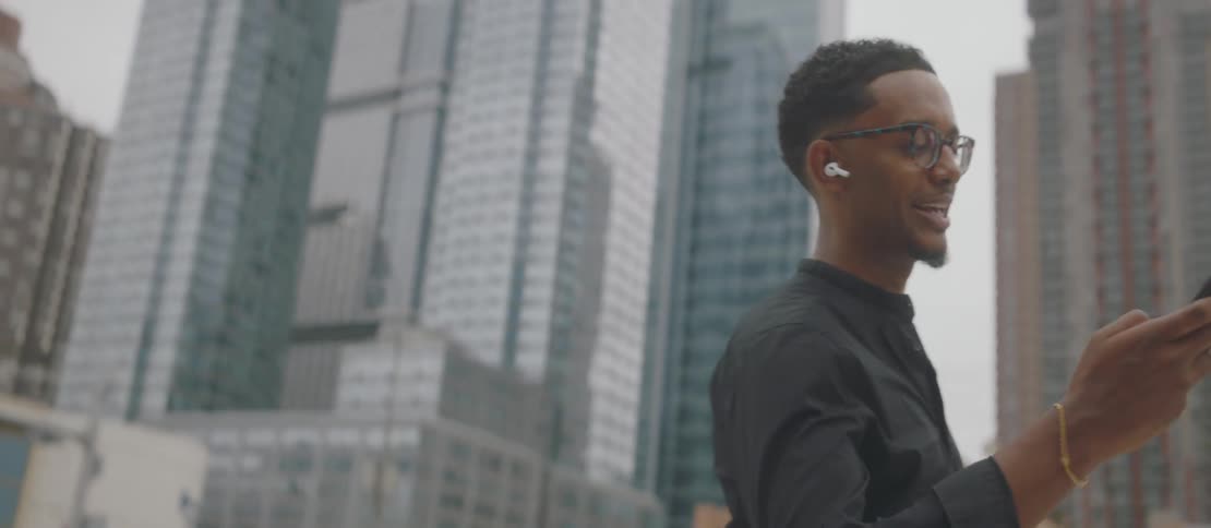 A young man wearing earbuds and reading glasses is walking and then looking at his cell phone and behind him there are several skyscrapers