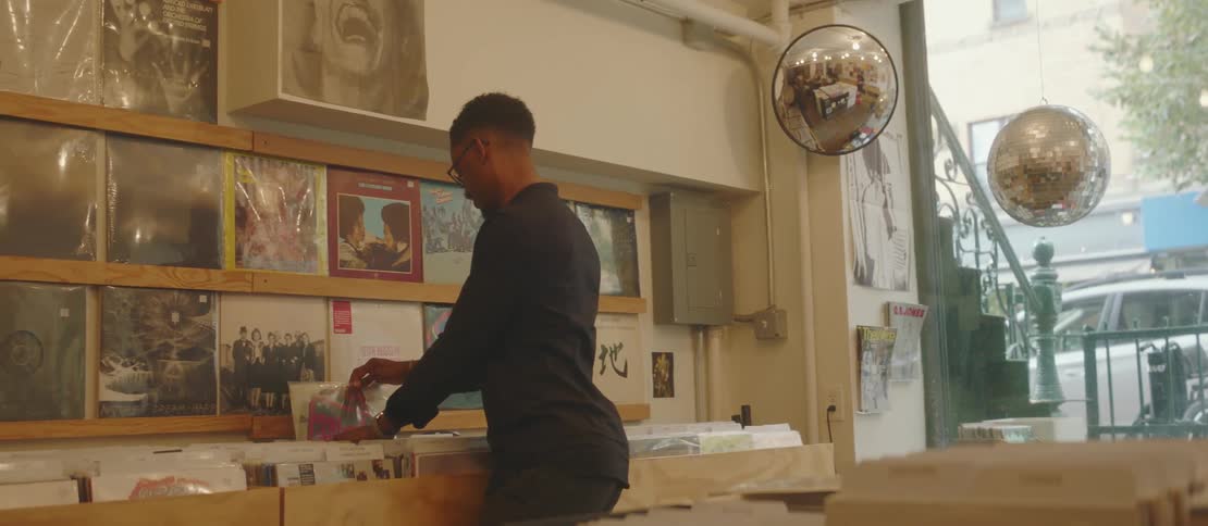 A black male looks through records at a store as a pedestrian passes by outside