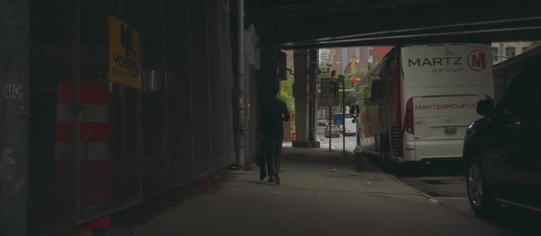A man walking on a sidewalk passing under an overpass looking at his smart phone while holding a bag