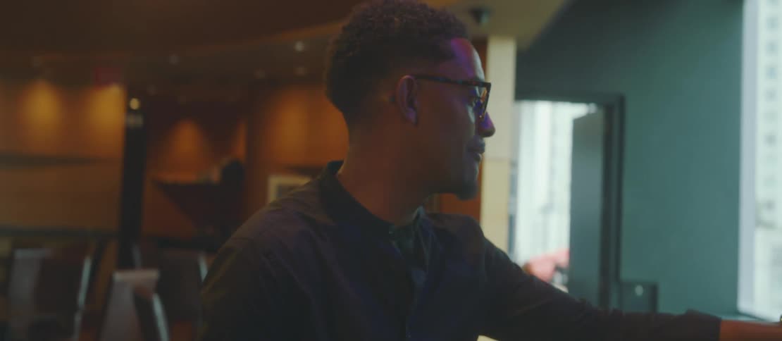 A man is sitting and wearing reading glasses while talking and looking at a cell phone that has an application open and on a surface of a piece of furniture